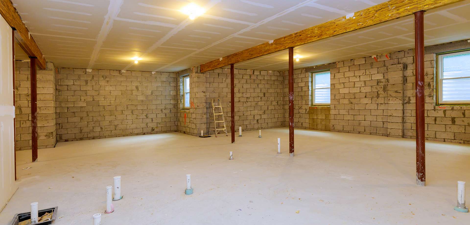 Unfinished basement with concrete floor, exposed cinder block walls, and ceiling joists—ideal for basement remodeling. Several plumbing pipes protrude from the ground. Four red steel columns, two windows, a wooden ladder, and bright overhead lighting are present.