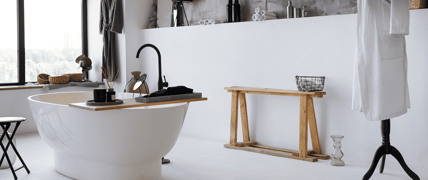 Modern minimalist bathroom remodeling features a freestanding white tub, black faucet, and tray with bath items. Towel and robes hang nearby; a wooden bench, wire basket, and toiletries rest on a platform shelf. Large window lets in natural light.