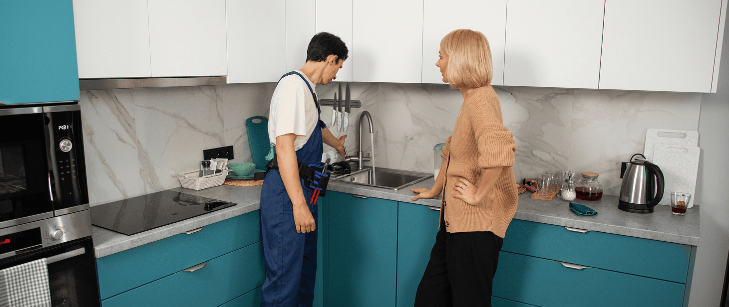 A plumber in blue overalls works under a kitchen sink while a woman with short blonde hair, wearing a brown sweater and black pants, watches—perhaps discussing her recent kitchen renovation with teal cabinets and modern appliances.