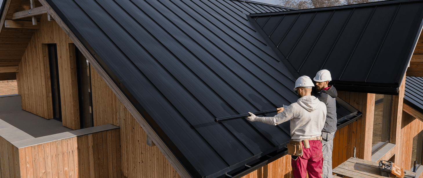 Two workers in hard hats stand on scaffolding, installing or inspecting a modern, dark metal roof on a wooden house. One holds a tool along a seam, likely involved in roof repair or replacement. The sky is clear and bare trees surround the house.