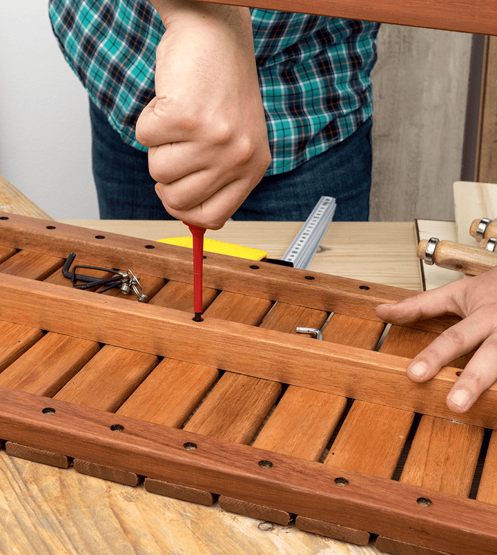 A person in a plaid shirt uses a red-handled screwdriver to fasten a screw into a wooden slatted surface, possibly assembling patios or decks. Other tools, screws, and metal components are visible nearby on a workbench, suggesting DIY woodworking.