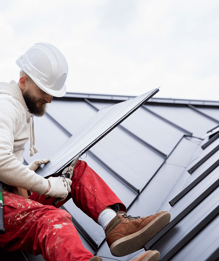 A construction worker in a white hard hat, hoodie, red pants, and brown boots installs a roof panel during a Roof Replacement on a modern dark gray metal roof. Seated, he adjusts the panel with a tool under a cloudy sky.