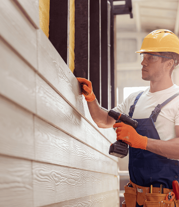 A construction worker wearing a yellow hard hat, orange gloves, and blue overalls uses a power drill for siding installation on a building. Focused, the worker holds a wooden plank in place as insulation is visible between wall beams.