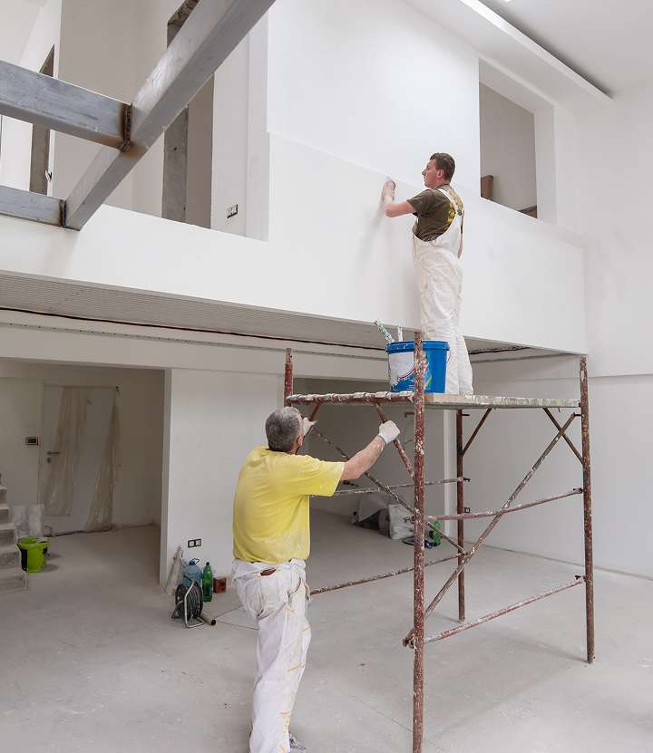 Two painters work inside a large, unfinished white room, part of a basement remodeling project. One paints a high wall from a scaffold while the other handles a lower section. Paint supplies and plastic coverings are scattered around the clean, under-construction space.