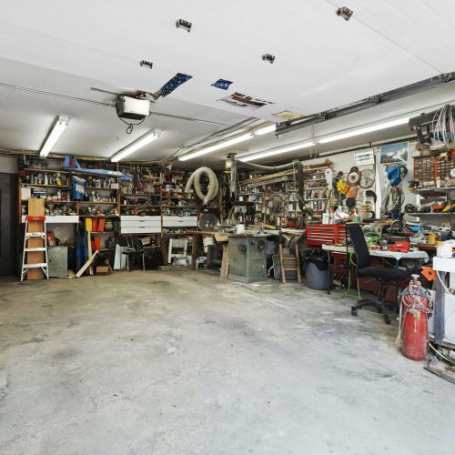 A cluttered garage workshop with various tools, equipment, and supplies on shelves and workbenches along the walls. The concrete floor is partially clear, and overhead lights illuminate the space.