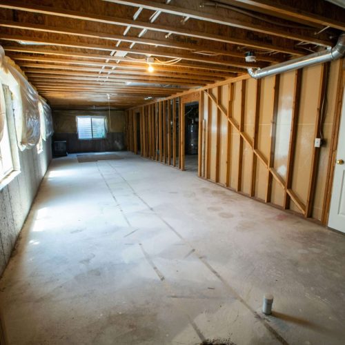 Unfinished basement with exposed wooden beams, concrete floor, and plastic sheeting on walls. A white door is on the right, and a small window is at the far end, letting in natural light.