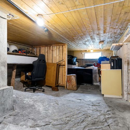 A basement room with a low wooden ceiling, rough stone floor, office chair at a cluttered desk, storage shelves, a bed, and various items stored along the walls.