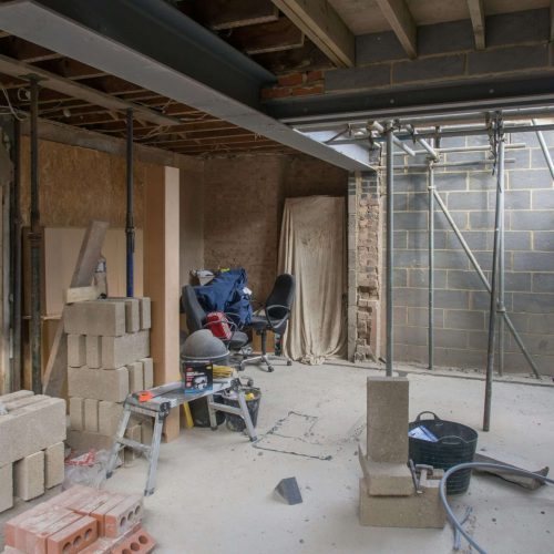Interior of a building under construction with exposed beams, stacked concrete blocks, scattered tools, a ladder, a black office chair, and unfinished walls. Natural light enters from the left side.