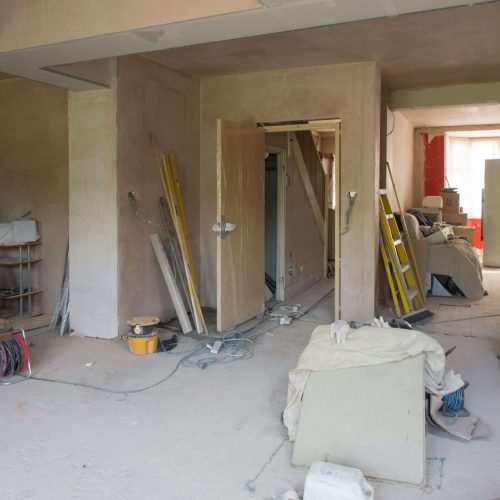 A room under renovation with unfinished walls, tools, ladders, cables, and construction materials scattered on the dusty floor. Furniture is pushed aside and covered, and natural light enters through a window in the background.