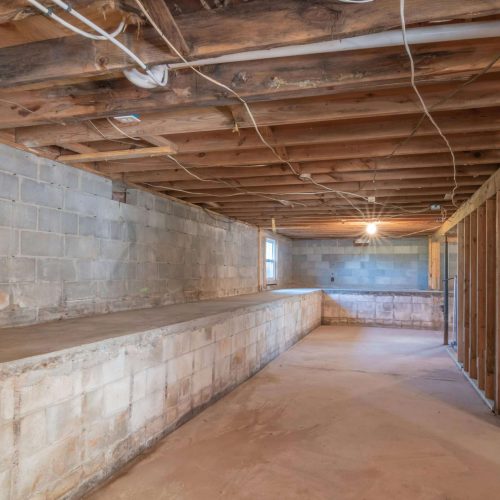 Unfinished basement with exposed wood beams and ceiling wires, cinder block walls, and a concrete floor. A raised concrete platform runs along one side, and stairs are visible in the corner. A single light bulb hangs from the ceiling.