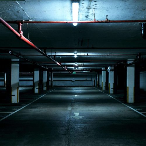 Dimly lit underground parking garage with concrete pillars, overhead pipes, and white arrows painted on the floor. The garage is mostly empty, and fluorescent lights cast a bluish tone across the space.