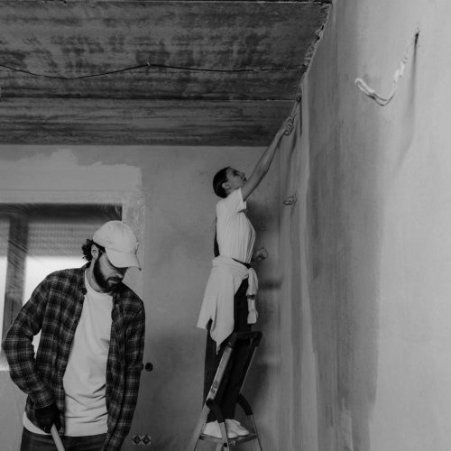 Two people are renovating a room; one stands on a ladder painting a wall while the other stands nearby, looking down and holding a tool. The room appears unfinished, with exposed wiring and an unpainted ceiling.