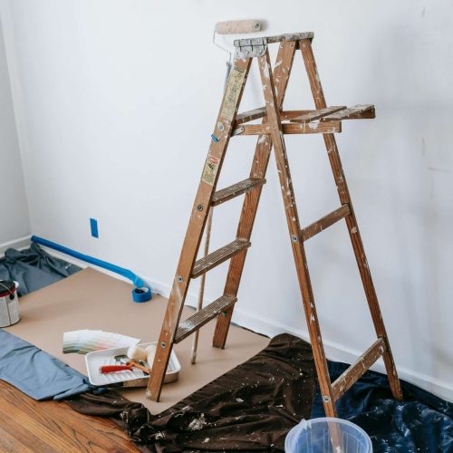 A wooden ladder stands in a room being prepared for painting, with paint cans, brushes, and rollers on a tarp-covered floor near a white wall. Blue painter’s tape lines the baseboard.