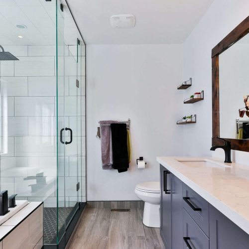 Modern bathroom with glass-enclosed shower, white bathtub, toilet, double-sink vanity with white countertop, large wood-framed mirror, and vase with dried flowers; shelves with toiletries on the wall.