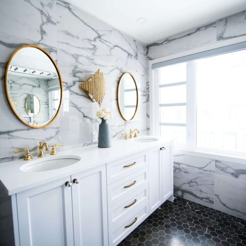 Modern bathroom with gray marble walls, a double white vanity with gold faucets, two round mirrors with gold frames, a gold wall sconce, a vase with flowers, and a large window letting in natural light.