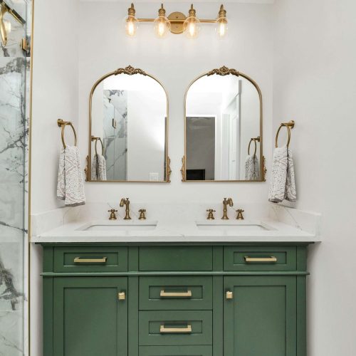 A bathroom features a green double-sink vanity with gold hardware, two arched mirrors, gold faucets, wall-mounted lights above each mirror, and marble tile seen on the left side of the image.