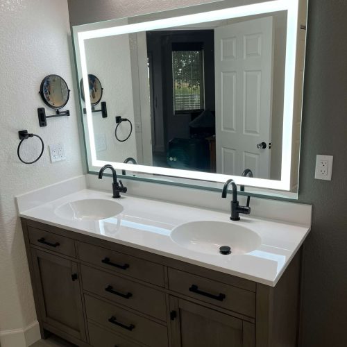A modern bathroom vanity with two sinks, black faucets, a large backlit mirror, wall-mounted lights above, and dual round magnifying mirrors on the wall. The vanity has multiple drawers and cabinets below.