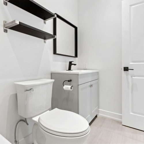 A modern bathroom with a white toilet, a vanity with a black faucet, a rectangular mirror, black wall shelves, and a white door. The walls and floor are light-colored.