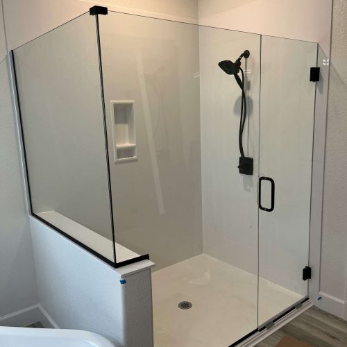 A corner shower with clear glass panels and a black frame features a black showerhead and handle, a built-in wall niche, and a white shower base. Tools and materials are on the floor nearby, indicating ongoing installation.
