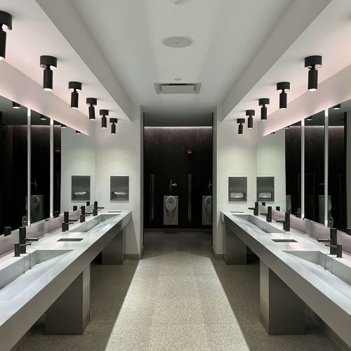 A modern public restroom with long sinks, wall-mounted faucets, mirrors, and automatic hand dryers on both sides. Two urinals are visible at the far end. The lighting is bright and evenly distributed.