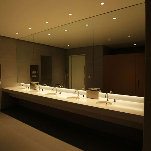 A modern public restroom with a long white countertop featuring four sinks, soap dispensers, a large wall mirror, gray tiled walls, and recessed ceiling lights. A door and stalls are visible in the background.
