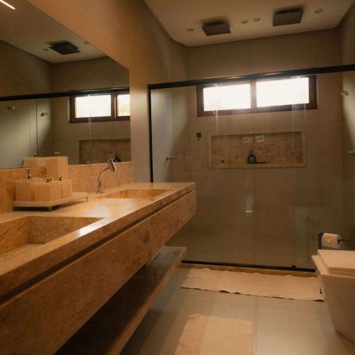 A modern bathroom with a beige stone double sink, large wall mirror, glass-enclosed shower, rectangular toilet, and soft lighting from a window above the shower area.