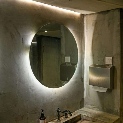 A circular backlit mirror above a concrete sink with a metal faucet, soap dispensers, and a metal paper towel dispenser mounted on the wall in a minimalist bathroom.