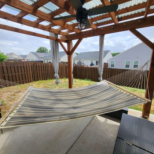 A striped hammock hangs under a wooden pergola with a ceiling fan and curtains in a fenced backyard. Nearby, there is a black outdoor bench on a concrete patio. Houses are visible in the background.