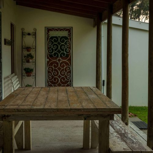 A rustic wooden table with benches sits on a covered porch next to a white house. There is a decorative metal door and a plant stand with potted plants against the wall. Grass and trees are visible in the background.