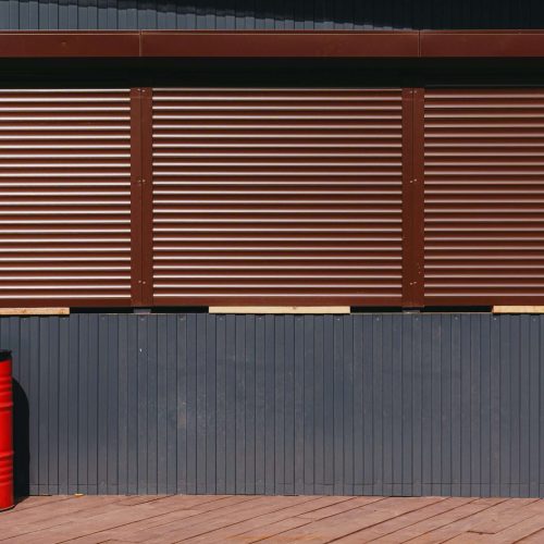 A red metal barrel stands on a wooden deck in front of a dark blue corrugated wall with three large closed brown metal shutters.
