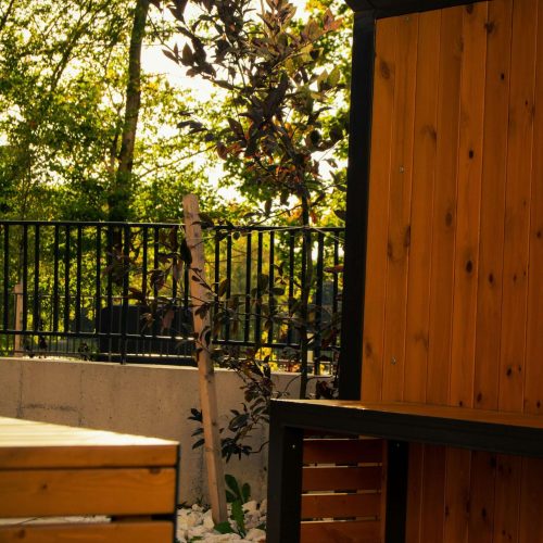 A wooden outdoor seating area with a bench and wall, next to a black metal fence, some greenery, and a tree in the background. Sunlight filters through the trees, casting shadows.