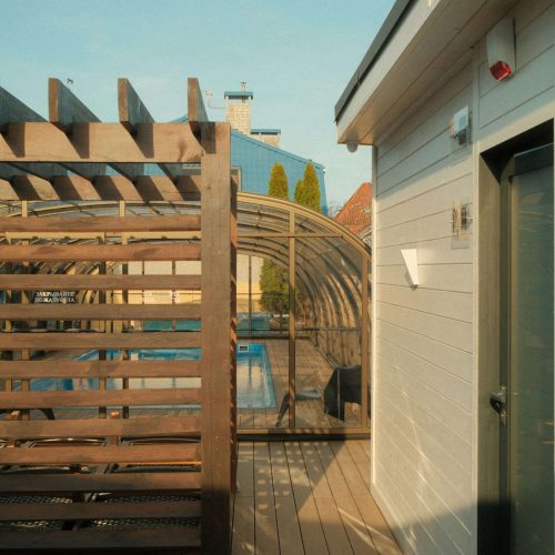 A wooden deck area with a pergola and a glass-enclosed pool in the background. The scene is lit by sunlight, with a modern white building on the right and a clear sky overhead.