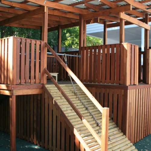 A wooden playground structure with vertical slats, an angled ramp made of logs, and a metal roof. The structure is elevated with open and enclosed sections, and surrounded by trees and a building in the background.