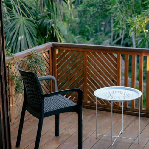 A black plastic chair and a small round white table sit on a wooden balcony with a railing, surrounded by green foliage and trees in the background.