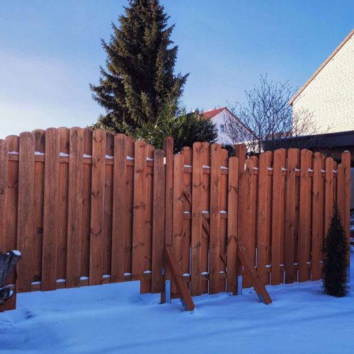 A wooden fence with damaged panels leans inward against a backdrop of snow-covered ground. A tree and residential houses are visible in the background under a clear blue sky.
