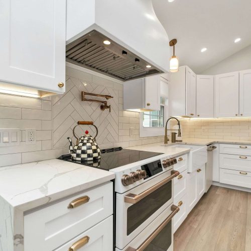 Modern kitchen with white cabinets, marble countertops, brass handles, a double oven, a black and white checkered kettle on the stove, under-cabinet lighting, and a light wood floor.