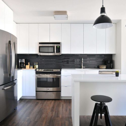A modern kitchen with white cabinets, stainless steel appliances, a black tile backsplash, dark wood flooring, a white island with two black stools, and two black pendant lights hanging above the island.