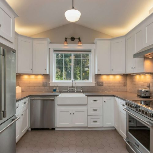 A modern kitchen with white cabinets, stainless steel appliances, a farmhouse sink under a window, beige subway tile backsplash, hexagonal floor tiles, and under-cabinet lighting.