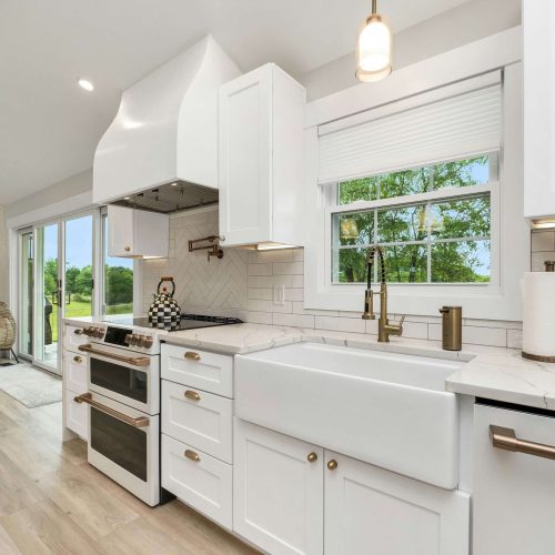 A modern kitchen with white cabinets, a farmhouse sink, stainless steel appliances, and gold hardware. A window above the sink shows green trees outside. The kitchen opens to a living area with a fireplace.