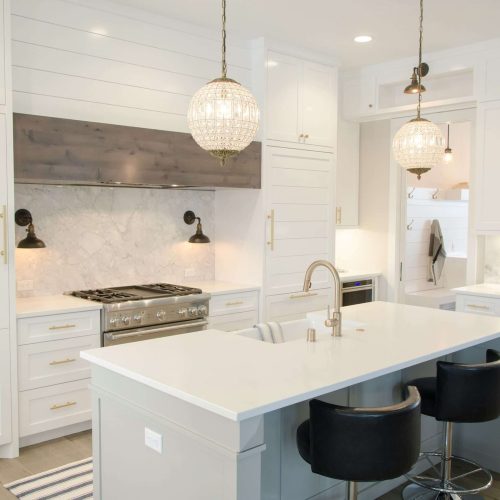 A modern kitchen with white cabinets, a marble backsplash, an island with a sink and two black chairs, stainless steel appliances, two pendant lights, and built-in shelves with dishes.