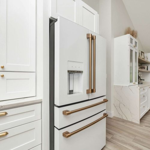 A modern kitchen with white cabinets and gold handles, a white refrigerator with gold handles and a water dispenser, light wood flooring, and a window letting in natural light.