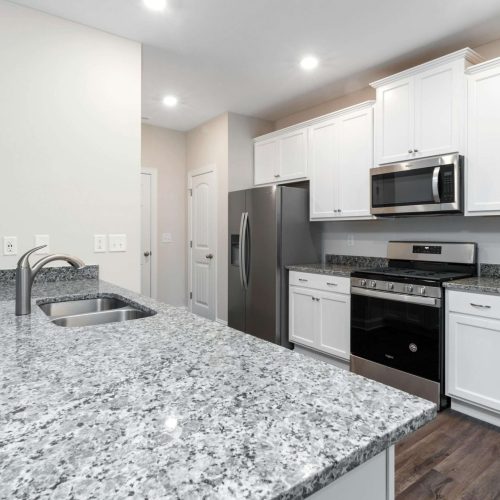 Modern kitchen with white cabinets, stainless steel appliances including a refrigerator, oven, and microwave, granite countertops, a double sink with a faucet, and wood-style flooring under bright ceiling lights.