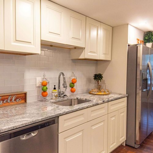 A modern kitchen with white cabinets, a stainless steel dishwasher and refrigerator, granite countertops, a sink with a faucet, and a backsplash made of white tiles. A dining area with a table and chairs is visible in the background.