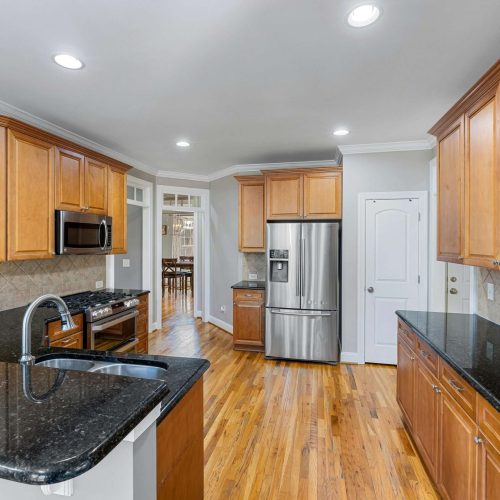 A modern kitchen with light wood cabinets, black granite countertops, stainless steel appliances including a fridge and microwave, wood flooring, and recessed lighting. A doorway leads to another room.