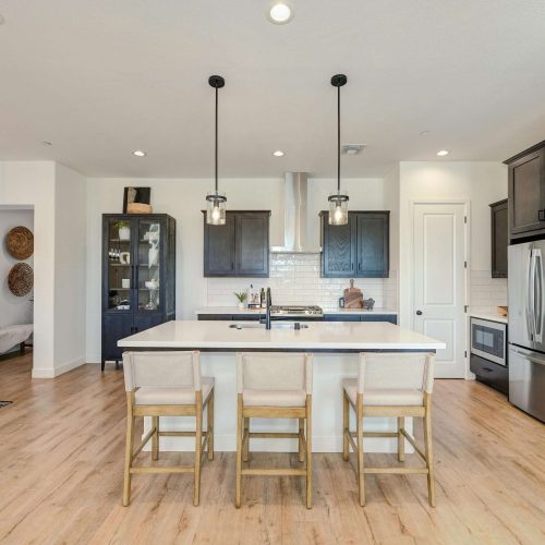 A modern kitchen with light wood floors, a large island with three beige chairs, dark cabinets, stainless steel appliances, pendant lights, and a white tile backsplash. A glass cabinet and decorative items are visible in the background.