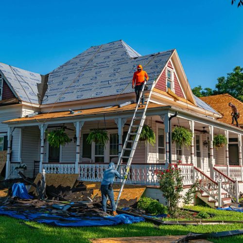 Workers are replacing the roof of a light-colored house, with roofing materials and debris scattered around the yard. One worker stands on a ladder, while others work on different parts of the roof under a clear blue sky.
