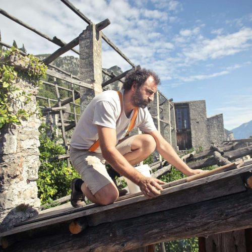 A man wearing a light shirt and shorts is kneeling on a wooden roof structure, working with his hands. The background shows stone walls, greenery, and a clear sky with scattered clouds.