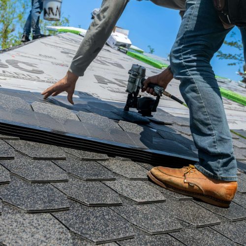 A person installing asphalt shingles on a roof using a nail gun, with roofing underlayment visible in the background on a sunny day.