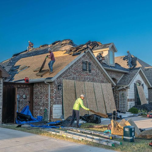Several workers are replacing the roof shingles on a brick house. Ladders, tools, and roofing materials are scattered on the driveway and yard. The sky is clear and blue.