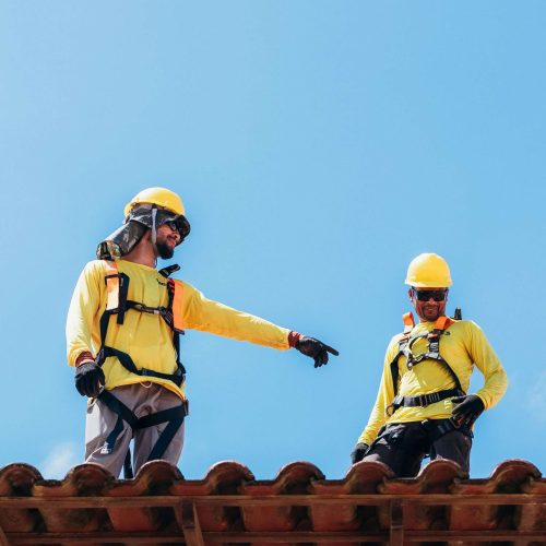 Two construction workers wearing yellow shirts, safety harnesses, and helmets stand on a tiled roof. One worker points while the other looks down. The sky is clear and blue in the background.